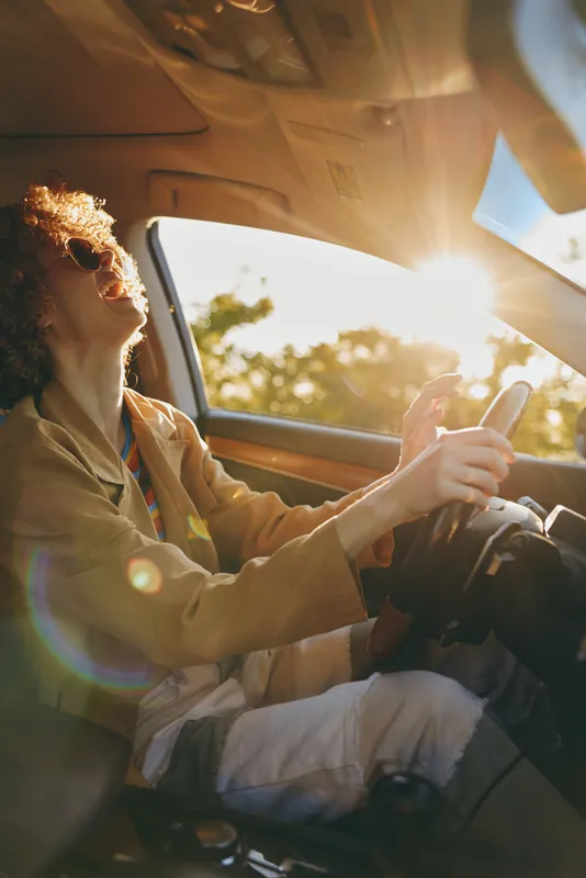 Smiling woman driving a car as sunlight bursts through the windshield, creating a warm retro film mood. Candid lifestyle shot with natural glow and relaxed road trip vibe.