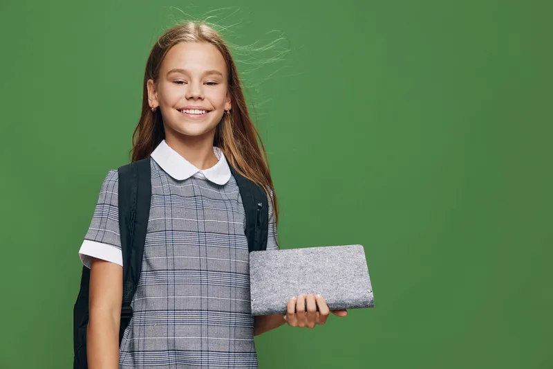 Smiling girl with closed eyes holding tablet standing with backpack in casual outfit on green background. Child showing digital device with happy expression in studio portrait.