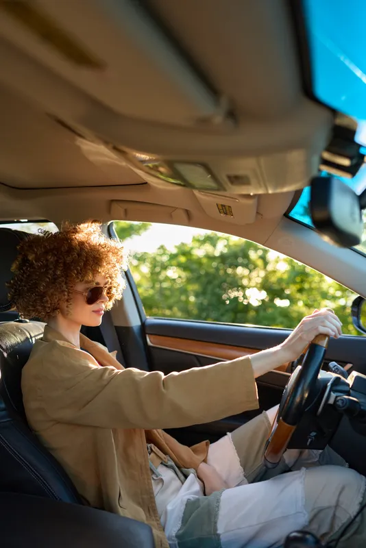 Cheerful woman driving a car, smiling with sunlit interior, casual outfit, relaxed posture, leafy greenery visible through window, vibrant colors, lifestyle moment with retro film warmth