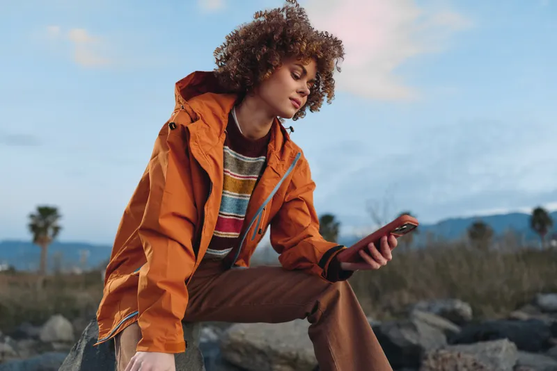 Woman lifestyle outdoors, sitting on rocks in orange jacket, looking at phone, curly hair, relaxed pose, bright daylight, distant mountains, casual street style, calm mood sunlit breeze