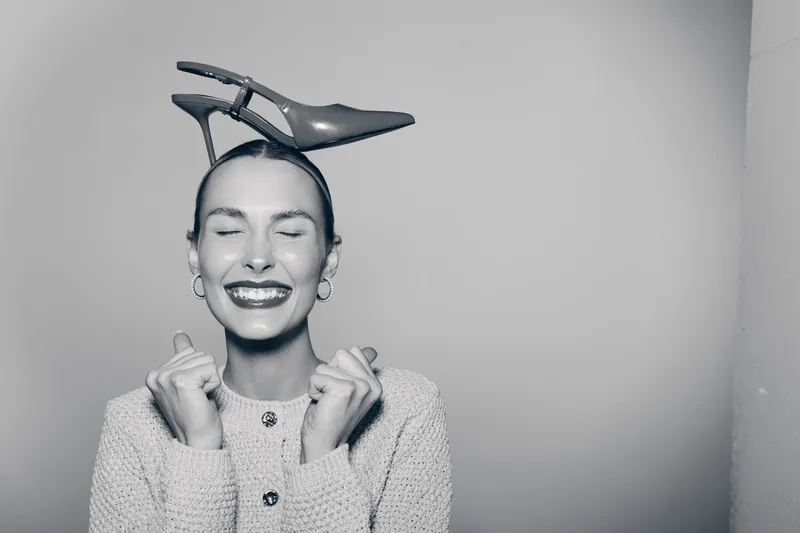 Happy caucasian woman with light skin smiles with closed eyes wearing high heel shoe on head, posing with clenched fists in monochrome studio. Concept of joy and fashion fun.