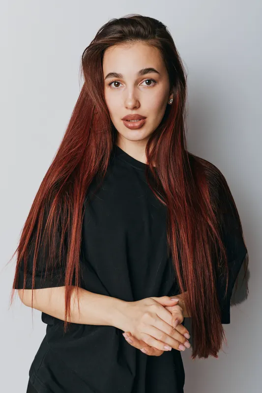 Young woman with long red hair and natural makeup wearing black shirt standing against white background with hands clasped expressing calm and confidence in studio portrait.