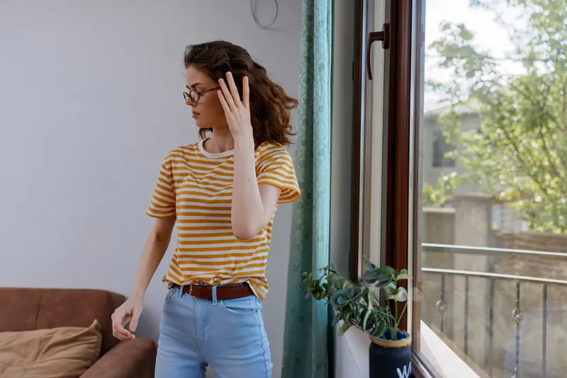 A woman with curly hair in a striped shirt stands in a bright home interior, adjusting her glasses, while looking toward a sunny window. Concept for everyday life and self reflection.