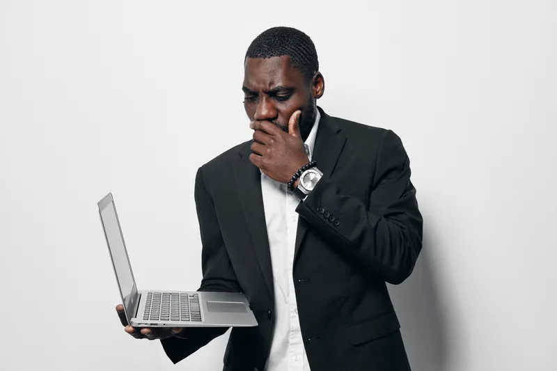 Black man with dark skin in formal suit holding laptop and looking thoughtful with hand on chin against white background. Business professional, technology and concentration concept.