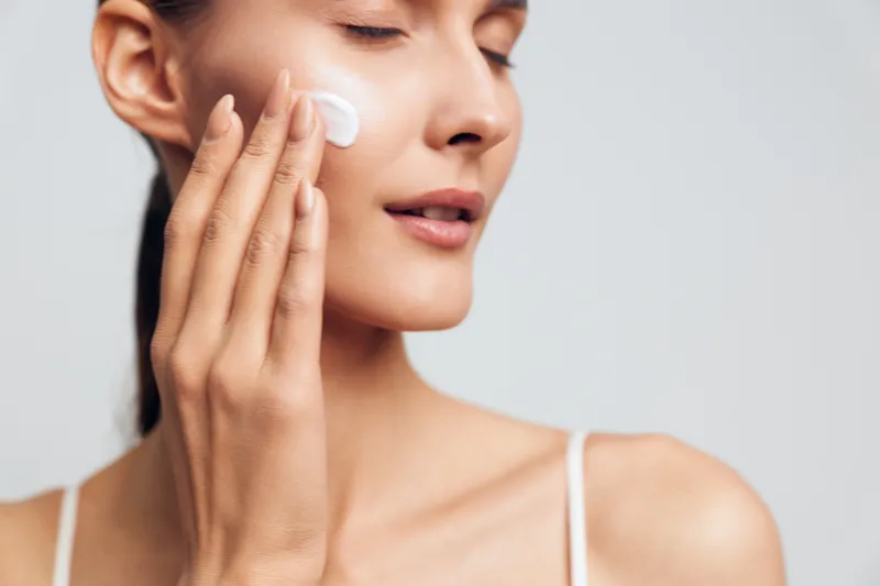 A young woman with clear skin applies moisturizing cream to her cheek with her fingers, eyes closed in a calm neutral background. Concept for skincare and beauty routine.