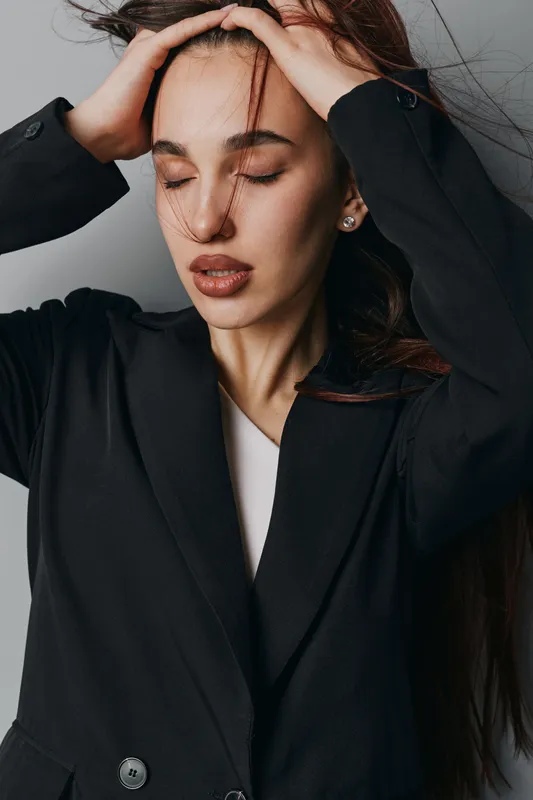 Young woman with long hair wearing black blazer posing with closed eyes and hands on head against gray background. Fashion, beauty, portrait, casual style and natural look.