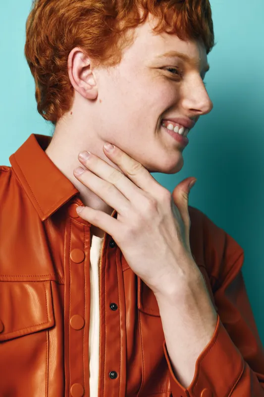 Young man with red hair wearing an orange leather jacket smiling and touching his neck against blue background, casual fashion portrait with natural expression and side profile.