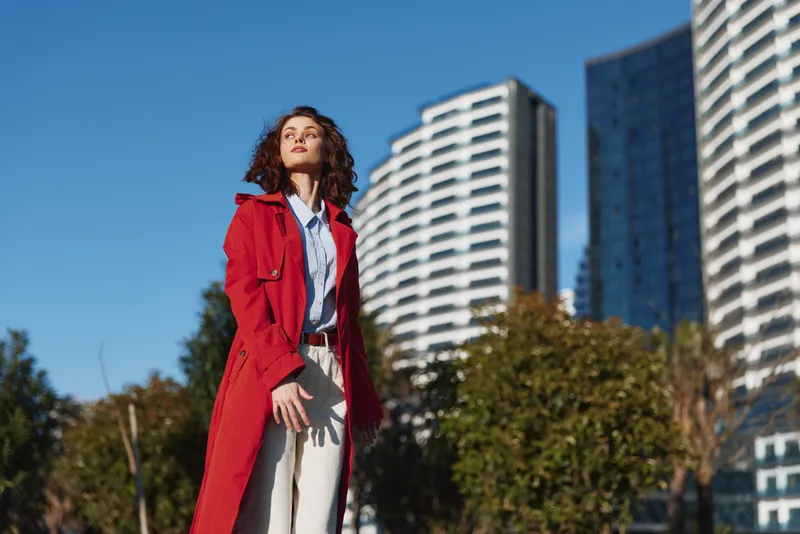A young woman with curly hair stands outdoors in a bright urban setting wearing a red trench coat, posing confidently against modern high rises.