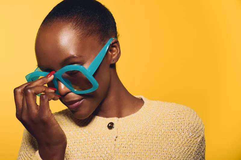 Portrait of a young African woman with dark skin wearing oversized blue sunglasses and a textured beige sweater against a bright yellow background. Concept of fashion, style, and confidence.