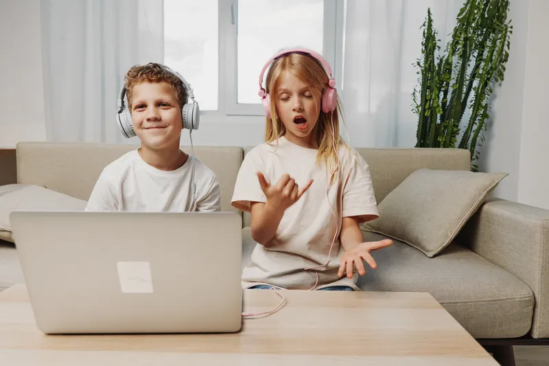 Two kids with headphones using laptop on sofa, boy smiling and girl singing with expressive face in bright living room.
