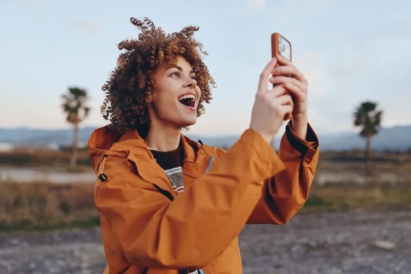 Woman lifestyle, outdoors selfie with smartphone, laughing and smiling in orange jacket, curly hair woman holding phone, travel portrait on road with palms and distant mountains at sunset