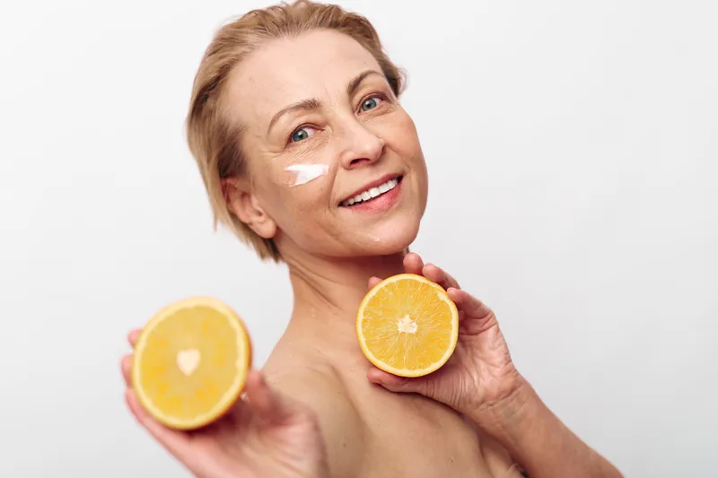 Smiling mature woman holding two orange halves near face with skincare cream on cheek. Concept of natural beauty, vitamin c, healthy skin, wellness, and age positive self care.