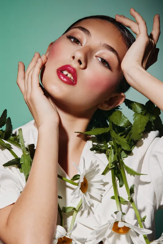 Young woman with glossy red lips and glowing skin poses with fresh daisies against soft green backdrop. Creative beauty and fashion portrait emphasizing elegance and floral freshness.