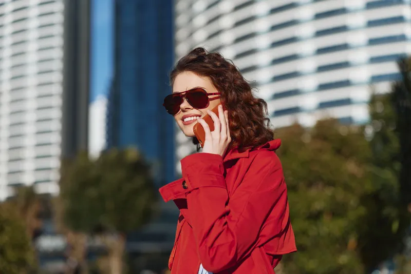 Young woman in a red jacket talks on her phone in a sunlit urban park, confident smile, relaxed pose, modern buildings behind, clear blue sky.