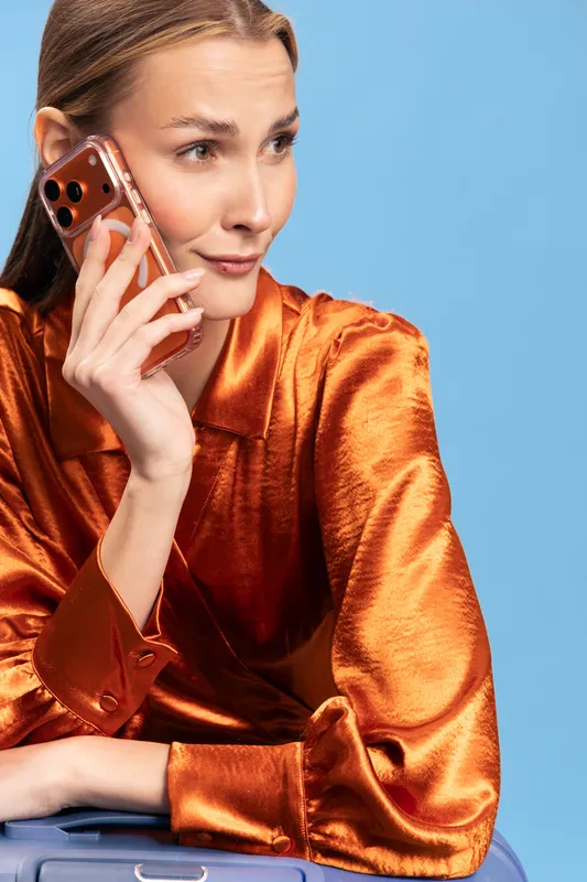 Young woman in orange satin blouse talks on smartphone with slight smile, leaning on suitcase against blue background. Concept of travel, communication, and fashion.