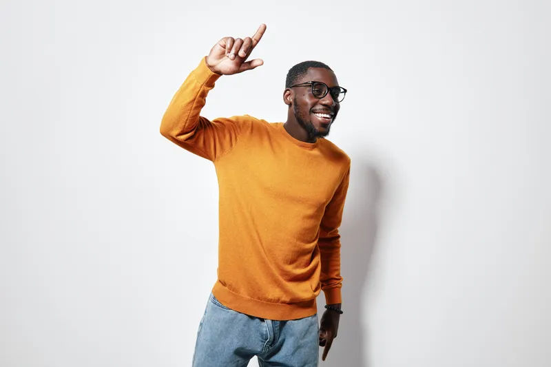 African American man with dark skin wearing glasses and orange sweater smiling and pointing up with finger against white background in casual style.