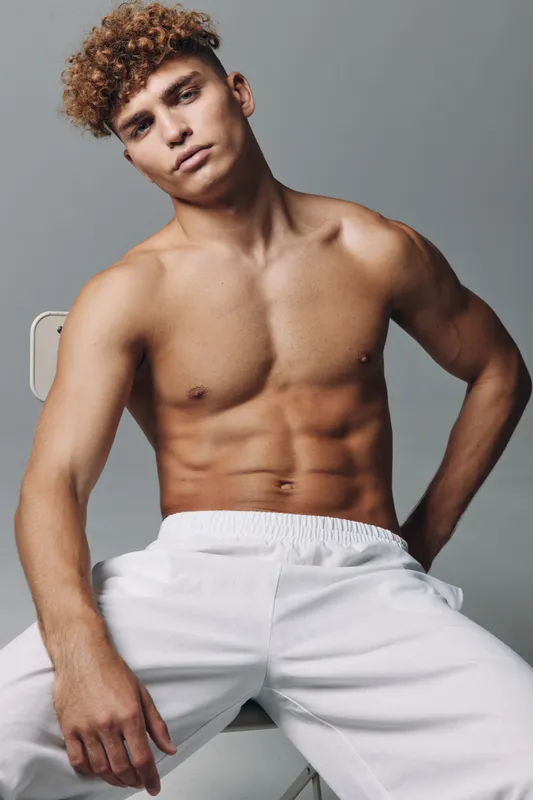 Muscular young man with curly hair posing shirtless with white pants indoors against gray background. Athletic body, defined muscles, and confident expression showing fitness and health.