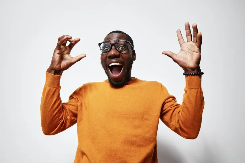 African American man with dark skin wearing glasses and orange sweater expressing surprise and excitement with open mouth and raised hands on white background.