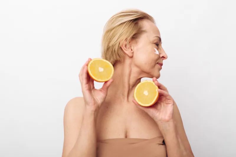 Mature woman with short blonde hair holds two halves of grapefruit near face with cream dots on cheek. Concept of natural skincare, healthy lifestyle, and beauty treatment at home.