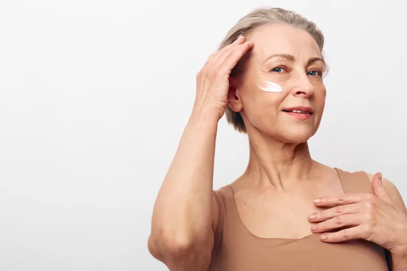 Mature woman with short hair applies white cream on face and neck, smiling gently against white background. Concept of skincare, aging, self-care, and beauty routine for older adults.