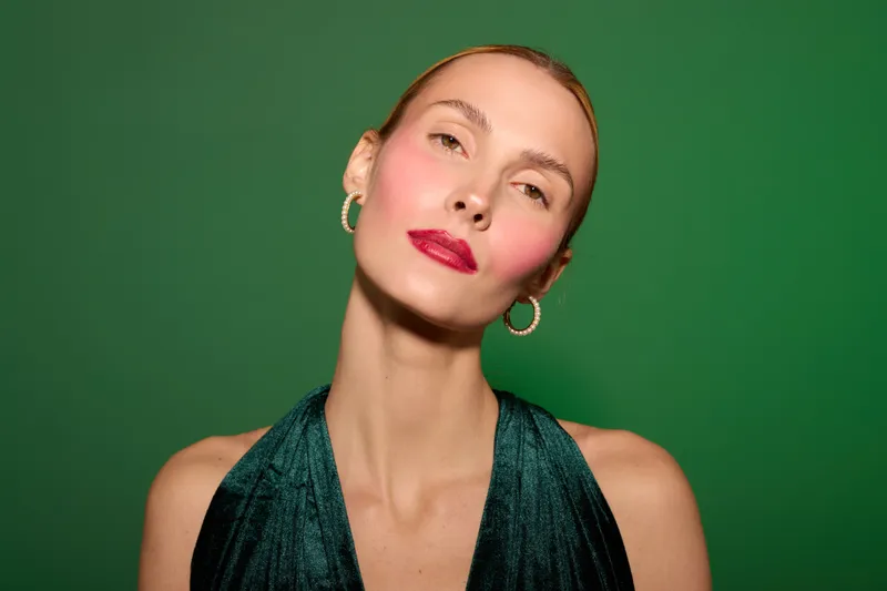Portrait of a confident woman with red lipstick and hoop earrings wearing a dark green dress against a green background. Concept of beauty, fashion, and elegance in studio lighting.