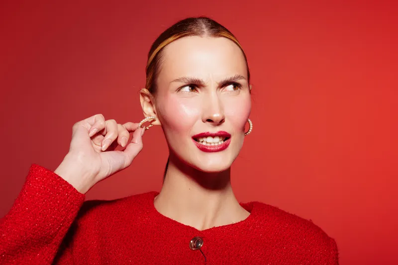 Young woman in red sweater pulls ear with finger, showing irritated expression on red background. Concept of discomfort, annoyance, and personal reaction to pain or noise.