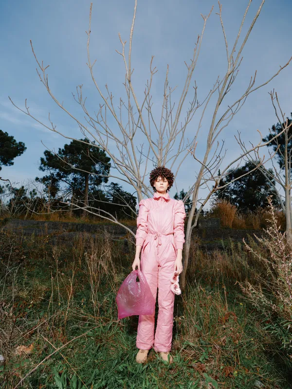 Fashion model in a pink jumpsuit stands beside a bare tree in an open field, holding a matching pink bag for a bold outdoor portrait with bold stance.