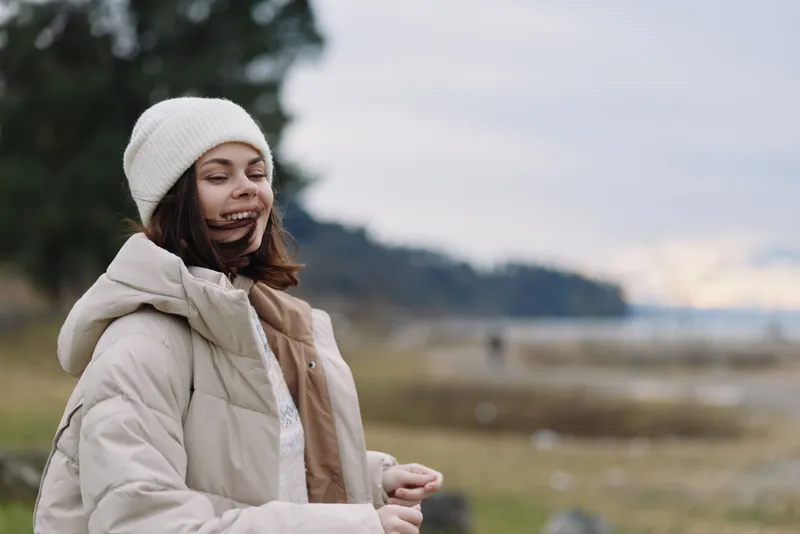 Woman in a puffer coat and knit hat enjoying a windy outdoor scene by a beach and shoreline with soft clouds and distant hills, conveying calm and freedom in nature