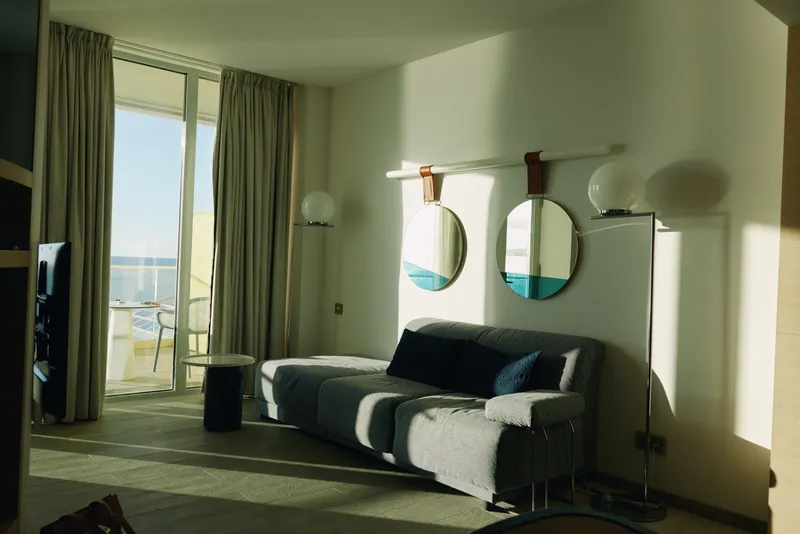 Bright hotel living room with a modern sofa, round mirrors, and large windows bathing the seating area in warm sunlight and offering a balcony view.