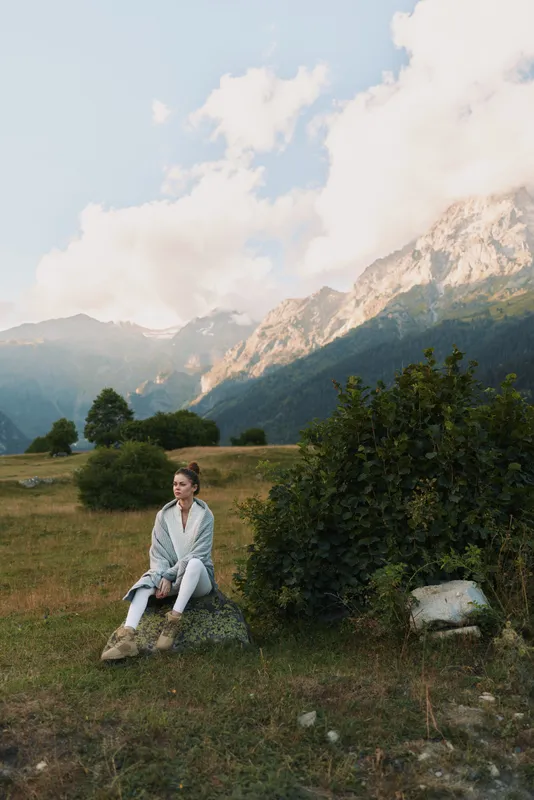 Woman lifestyle sitting on rock in mountain meadow, casual outfit, thoughtful expression, natural light, scenic landscape, cloudy sky, peaceful atmosphere