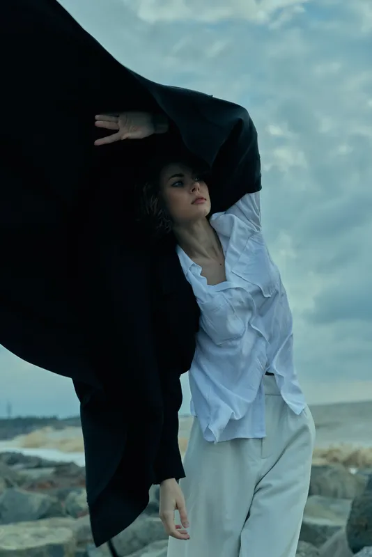 Moody scene of a woman on a windswept coastline, her cape draped over a dark fabric as she leans beneath stormy skies, with ocean rocks and surf in the background, creating drama