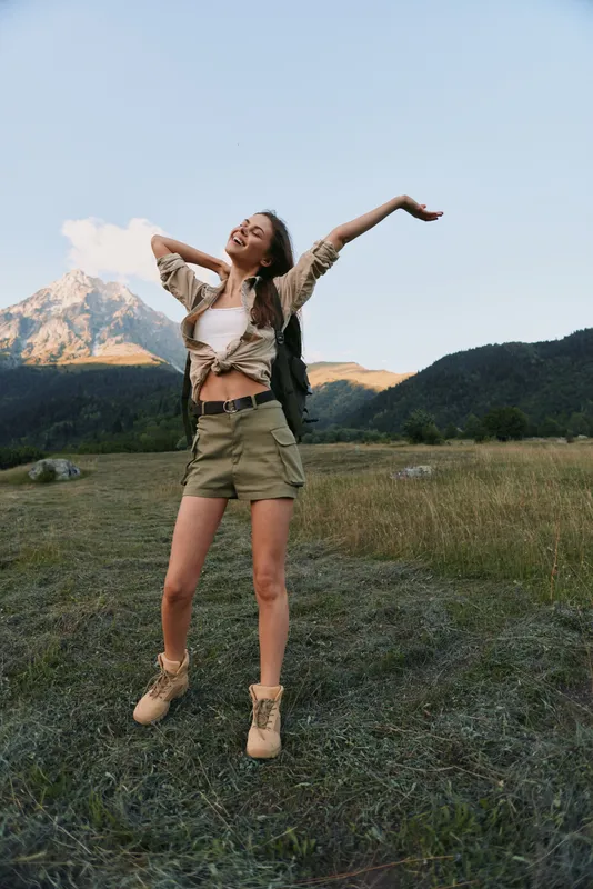 Young woman lifestyle outdoors stretching arms up enjoying fresh air in mountain field casual outfit sunny day natural beauty relaxed mood