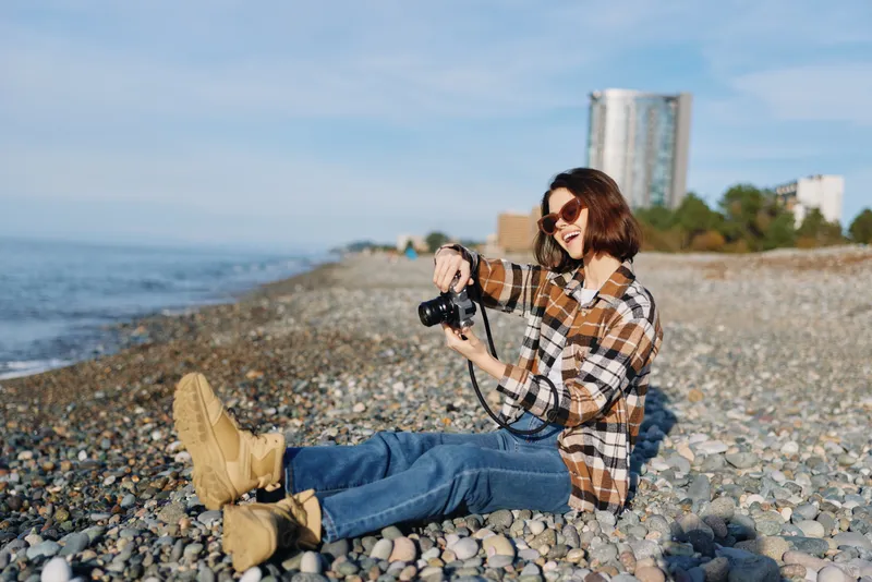 Beach photographer woman sits on pebbles by the sea, using a camera, enjoying leisure outdoors and sunlight, capturing candid coastal scenes with a relaxed fashion vibe and joyful energy.