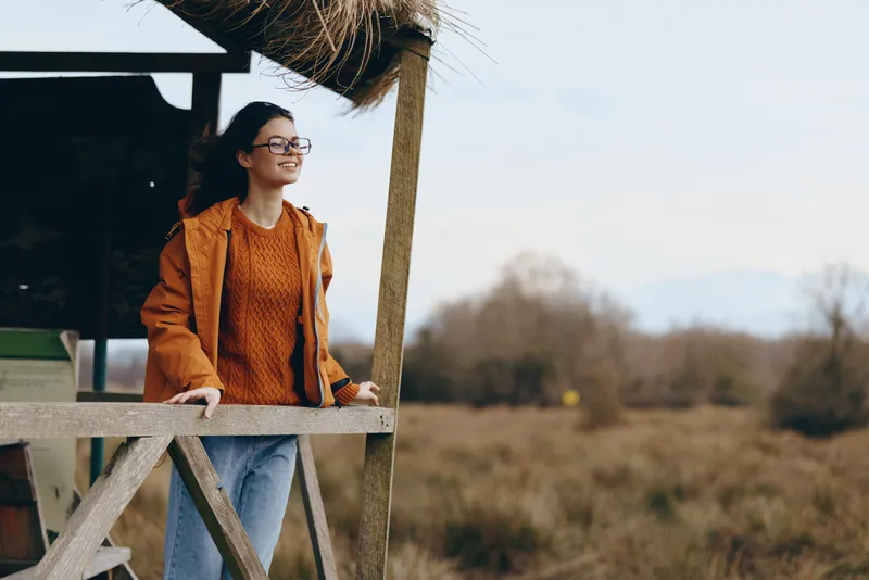Woman lifestyle outdoors in an autumn field, portrait with coat and hat, standing by wooden deck and rail, hand on support, gentle daylight, blurred trees and grass background, calm mood