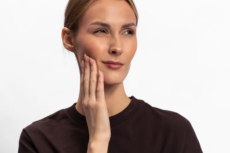 Young woman with clear skin touches her cheek with a worried expression on white background. Concept of toothache, facial pain, health problem, discomfort, and care.