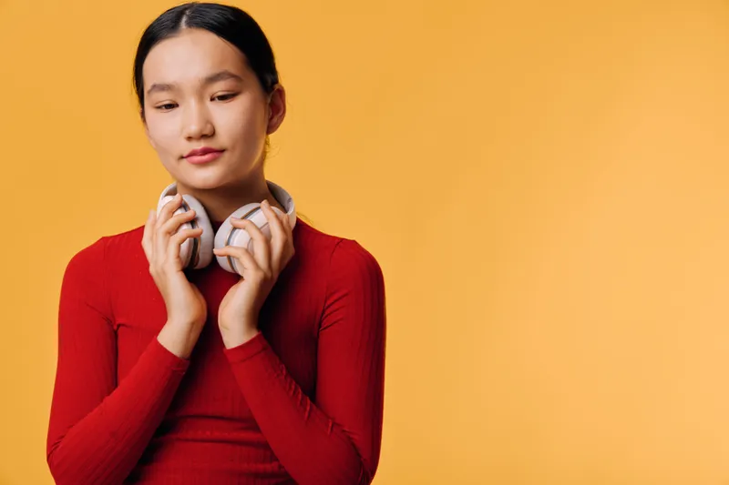 Young woman with headphones holding them near her neck in front of orange background. Casual style, thoughtful expression, and closeup portrait of female with long hair and red top.