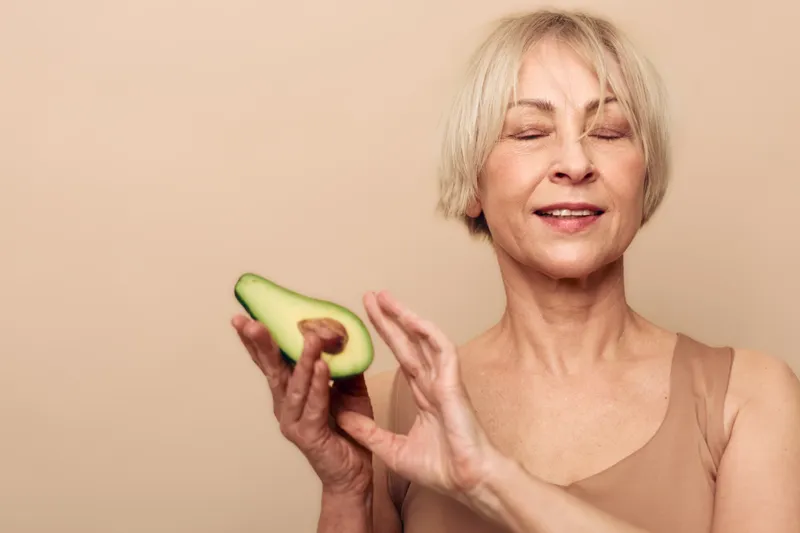 Happy mature woman with short hair holding fresh avocado half smiles with closed eyes on beige background. Concept of healthy lifestyle, nutrition, natural food, and wellness at any age.