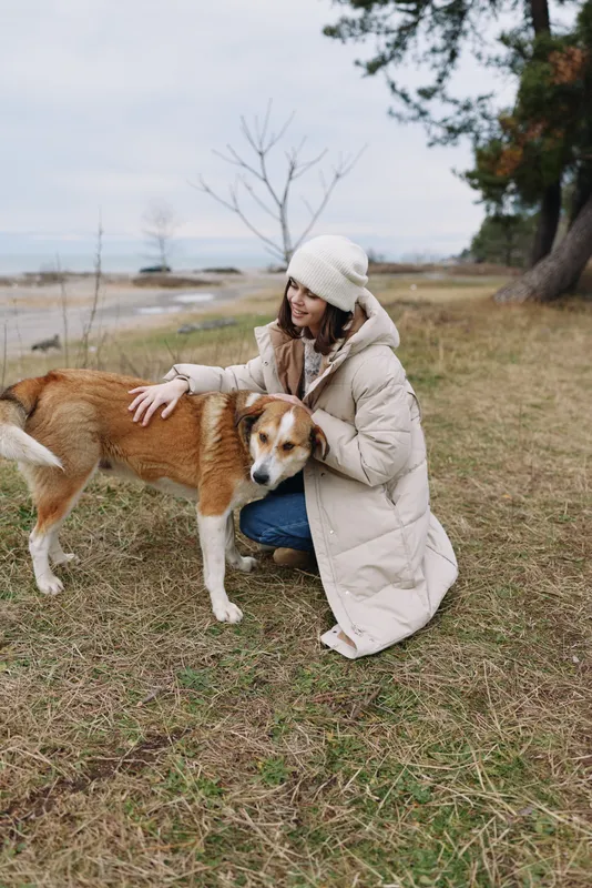 Woman in a warm coat and knit hat gently pets a dog in a park by a calm lake, autumn grass and trees in the background, a peaceful outdoor moment.