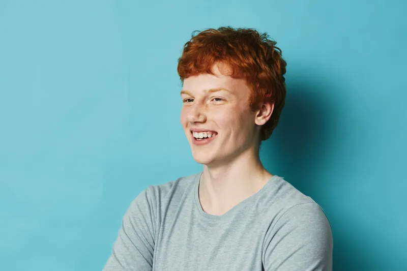 Young man with red hair and freckles smiling looking away wearing gray t shirt on blue background. Portrait of happy confident male with casual style and natural expression.