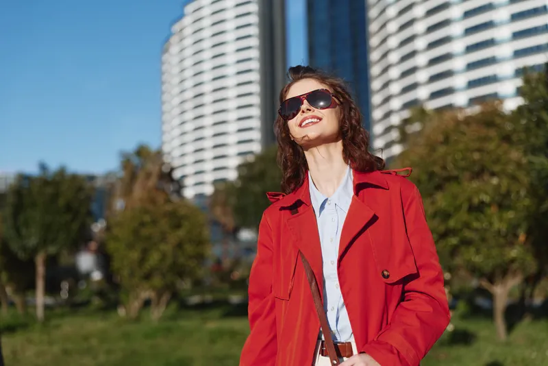 A young woman in a red coat and sunglasses stands in a sunny park with modern buildings behind, radiating confident, casual fashion.
