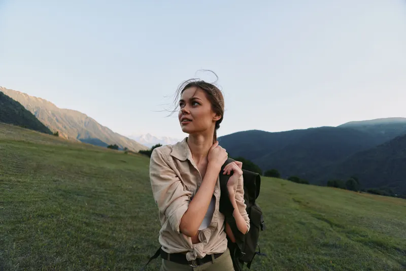Young woman traveler stands on green mountain meadow with backpack looks away thoughtful enjoying nature and fresh air during hiking trip in clear sky daylight.