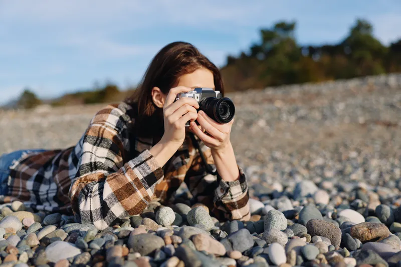 Photographer, outdoors, pebbles, beach, lens, candid, hobby, woman lying on a rocky shore and taking a photo with a camera under a clear blue sky.