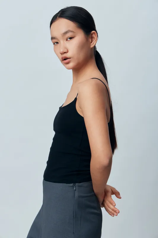 Young woman with long dark hair wearing black tank top and gray skirt posing with hands behind back against plain light background in studio portrait. Serious expression and casual style.