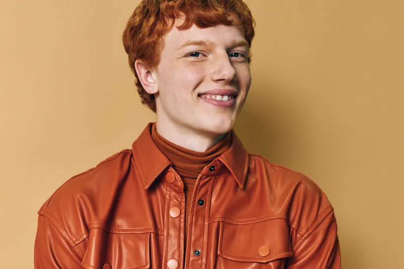 Young man with red hair wearing orange shirt and turtleneck smiling confident against beige background, portrait with casual style and positive expression.