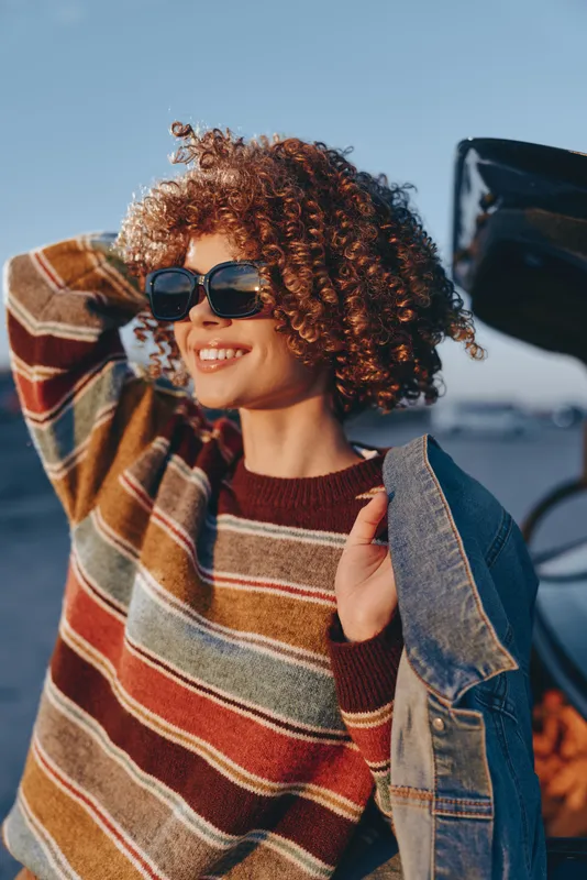 Woman lifestyle outdoors, smile behind sunglasses, curly hair, striped sweater, denim jacket, sunny street light, hand touching lapel, casual fashion mood, soft blue sky