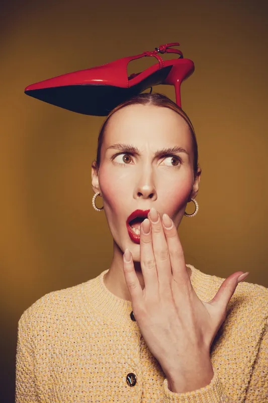 Surprised woman with red lipstick and hoop earrings holds hand near mouth, balancing red high heel shoe on head. Concept of fashion, surprise, and unusual style in studio lighting.