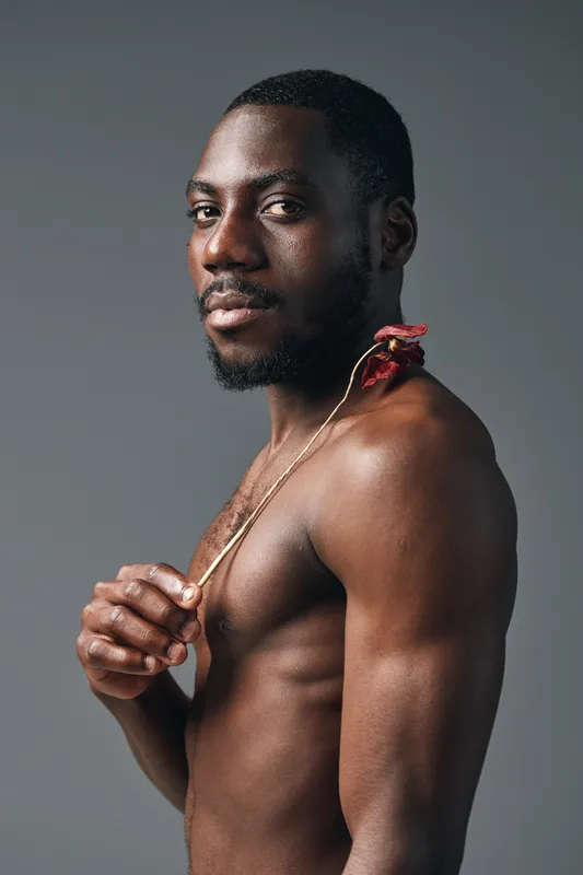 African man with dark skin and beard posing shirtless in creative conceptual style holding a flower on shoulder against gray background in studio portrait.