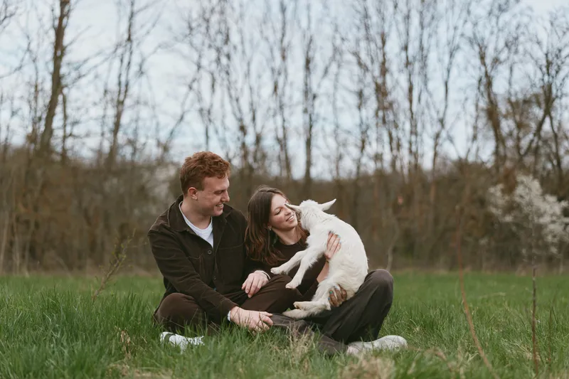 Candid outdoor scene shows a couple with a goat kid in a green field, capturing lifestyle, affection, smiling faces, relaxed posture, field, nature, family, happiness, tenderness