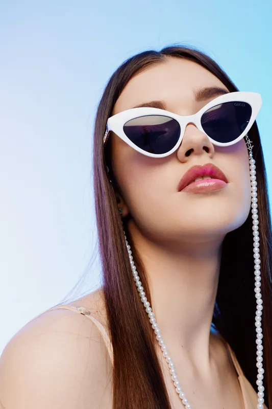 Young woman beauty fashion wearing white sunglasses with pearl chain looks confident against blue background. Close-up portrait with long hair and pink lips. Stylish concept.