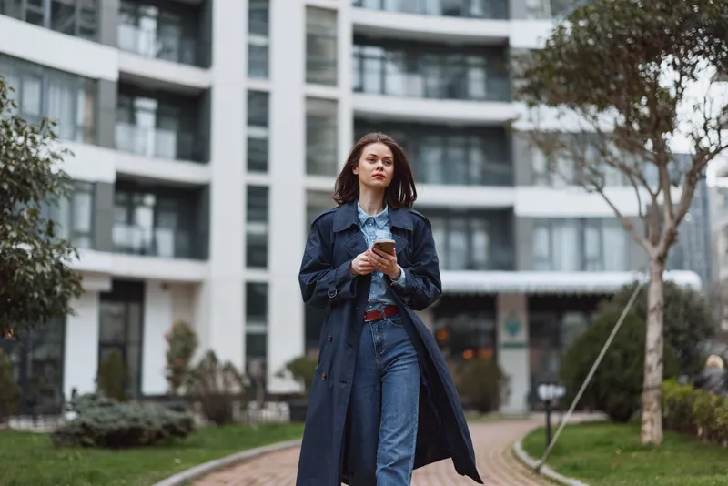 Young woman in denim trench and jeans walks along an urban campus path while looking at her phone, exuding confidence and casual street style.
